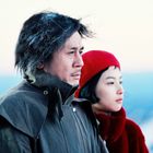 Ein Mann und eine Frau stehen nebeneinander, schauen ernst in die Ferne. Der Mann trägt dunkle Kleidung, die Frau trägt Rot.