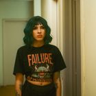 Text: "FAILURE" auf rotem T-Shirt mit Dämonengesicht. Frau mit grünen Haaren und Tattoos steht im Flur.
