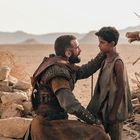 Ein Mann in Rüstung kniet in einer Wüstenlandschaft vor einem Jungen in zerlumpter Kleidung. Im Hintergrund sind Berge.
