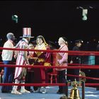 Zwei Boxer im Ring, einer trägt einen Hut in US-Farben, rote Seile, umgeben von Trainern und Schiedsrichtern.