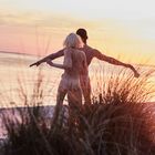 Zwei Menschen stehen am Strand bei Sonnenuntergang, sie haben die Arme ausgebreitet. Gräser im Vordergrund.