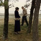 Zwei Personen stehen zwischen Bäumen auf einer Wiese, eine Frau links, ein Mann rechts in Gehrichtung. Im Hintergrund ein Haus.