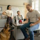 Eine Frau und zwei Männer in einem Büro, mit einem Schäferhund. Einer der Männer hält einen roten Ball in der Hand.
