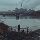 Eine graue, industrielle Landschaft mit rauchenden Fabriken, einem Fluss im Vordergrund und zwei Menschen, die am Ufer stehen.