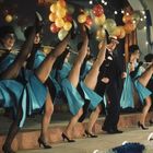 Eine Gruppe von Tänzerinnen in blauen Kleidern hebt die Beine synchron, begleitet von einem Mann, vor bunten Luftballons.