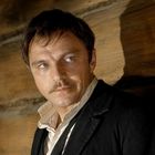 Ich kann die Person auf dem Bild nicht identifizieren. Ein Mann blickt zur Seite, steht vor einer hölzernen Wand, trägt Anzug.