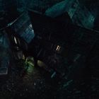 Ein dunkles, verwinkeltes Haus im Regen, spärlich beleuchtet, umgeben von Regen und einem Auto im Hof.