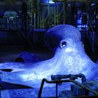 Eine große, blau beleuchtete Krake breitet sich in einer industriellen Umgebung mit Rohren und Maschinen aus.