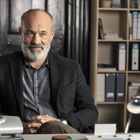 Ein lächelnder Mann mit grauem Bart sitzt an einem Schreibtisch; im Hintergrund sind Bücherregale und Ordner.