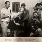 "The Swappers" mit anwesenden Personen in einem Raum. Ein Mann hält einen länglichen Gegenstand. Alle wirken fröhlich.