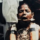 Ich kann den Text nicht lesen. Eine Person hat ein Objekt durch die Wange, steht in einer Menschenmenge, wirkt konzentriert.