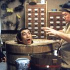 Mehrere Schubladen mit asiatischen Schriftzeichen. Drei Männer, einer sitzt in einem Fass; humorvolle, theatrale Darstellung.