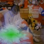 Ein Schild mit der Aufschrift „STOPPING 6 P.M.“. Eine Person sitzt auf einem Bett voller Spielzeug und Geld. Ein grüner Fleck verdeckt teilweise die Szene.