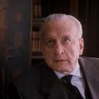 Ein älterer Mann mit Anzug sitzt in einem Raum mit Büchern im Hintergrund. Er hat graues Haar und schaut aufmerksam.