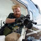 Eine Person mit rotem Bart zielt ernst aus einem Helikopter mit einem großen Gewehr.