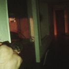 Ein dunkler Korridor mit rotem Licht. Links kniet eine Person, an die Wand gelehnt. Mysteriöse, düstere Atmosphäre.