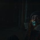 Eine Person mit Klebeband über dem Mund sitzt verängstigt in einem schwach beleuchteten Raum, trägt eine Mütze. Links oben: Licht.