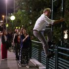 Eine Person klettert nachts über ein Brückengeländer, während mehrere Menschen besorgt zuschauen. Die Straße ist beleuchtet.