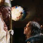 Ein Geistlicher mit gold-roben kniet vor einer Person in Rüstung. Im Hintergrund ein bunt leuchtendes Kirchenfenster.