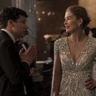 Zwei elegant gekleidete Personen, eine Frau in einem glänzenden Kleid, ein Mann im Anzug, stehen in einem festlichen Raum.