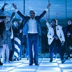 Eine Gruppe von Menschen tanzt fröhlich auf einem beleuchteten Boden in einem festlichen, blauen Licht.