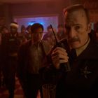 Ein Polizist, erkennbar an Sheriff-Abzeichen, spricht in ein Funkgerät. Mehrere Personen stehen im Hintergrund in einem Lokal.
