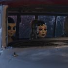 Vorne im Auto sitzen drei Personen mit unheimlichen Masken, darunter eine mit roten Lippen und geschminkten Augen.