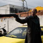 Ein Mann in einem Ledermantel hält ein Gewehr. Im Hintergrund steht ein gelbes Auto mit dem Schriftzug "Kamikoku".