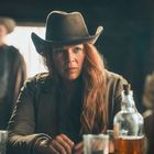 Keine sichtbaren Texte. Eine Frau mit Cowboyhut sitzt ernst an einem Tisch in einem düsteren Raum, mit Gläsern und Flasche.