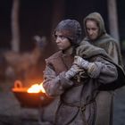 Eine Gruppe in mittelalterlicher Kleidung steht im Freien. Eine Person hält einen Stoff, im Hintergrund brennt ein Feuer.