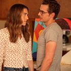 Zwei Personen, eine Frau mit langem Haar und ein Mann mit Brille, stehen sich nah gegenüber in einer Küche mit buntem Kunstwerk.