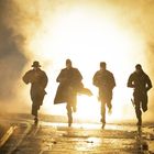 Vier Personen rennen in dunkler Kleidung durch eine neblige Szene, beleuchtet von einem intensiven Licht im Hintergrund.