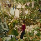 Eine Frau in grün, ein Kind und eine Figur in elegantem Anzug stehen auf einem hölzernen Balkon, Wäsche hängt über ihnen.