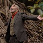 Ein älterer Mann in Anzug und rotem Schal breitet die Arme lächelnd aus, vor großen Baumwurzeln.