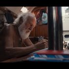 Ein älterer Mann mit weißem Bart sitzt in einem Holzkabinenraum und schreibt etwas mit einem Stift auf Papier.