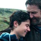 Zwei Personen, wahrscheinlich Vater und Sohn, umarmen sich lächelnd in einer grünen, hügeligen Landschaft.