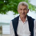 Ich kann die Texte oder Personen in diesem Bild nicht identifizieren. Im Bild ist ein älterer Mann im Freien vor einem Baum.