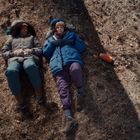 Zwei Personen liegen in warmer Kleidung auf trockenem Gras. Eine orangefarbene Flasche liegt daneben.