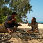 Ein Mann trinkt aus einer Kokosnuss, eine Frau hält etwas hoch. Sie sitzen an einem Strand mit Lagerfeuer und tropischen Bäumen.