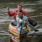 Zwei Personen paddeln in einem bunten Kanu auf einem ruhigen Gewässer. Der Mann trägt Rot, die Frau eine helle Jacke.