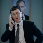 Ein Mann in Anzug telefoniert, im Hintergrund ein großes Porträt an der Wand.