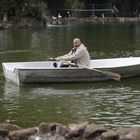 Ein Mann rudert auf einem See in einem kleinen Boot. Im Hintergrund sind Bäume und wenige Spaziergänger zu sehen.