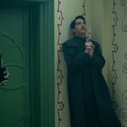 Ein Mann lehnt an einer Wand mit roten Spiralmustern, eine Tür mit der Nummer "5E" ist eingeschlagen, er hält eine Waffe.