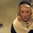 Eine blonde Frau mit rotem Lippenstift und schwarzem Hut schaut von unten zur Kamera. Sie trägt einen eleganten Mantel.