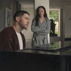 Ein Mann spielt Klavier, eine Frau steht daneben. Im Hintergrund sieht man eine Skulptur und Weinflaschen.