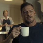 Ein Mann mit Bart hält eine Tasse, zwei Personen in Uniformen im Hintergrund, möglicherweise in einer Küche.