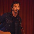 Ein Mann spielt Gitarre und singt in ein Mikrofon. Er trägt eine Jeansjacke und hat ein Pflaster auf der Stirn.
