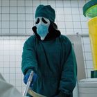 Der Text "THE STUFF" steht auf einem großen Behälter. Eine Person in OP-Kleidung mit Maske hält ein Gerät in der Hand.