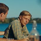 Es sind keine Texte im Bild. Drei Männer am Tisch vor einem See, sommerlich gekleidet. Einer trägt einen Strohhut.