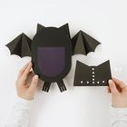 Eine kreative Fledermausbastelei aus schwarzem Papier mit zwei Händen. Origami-ähnliche Struktur und ausgeschnittene Details.